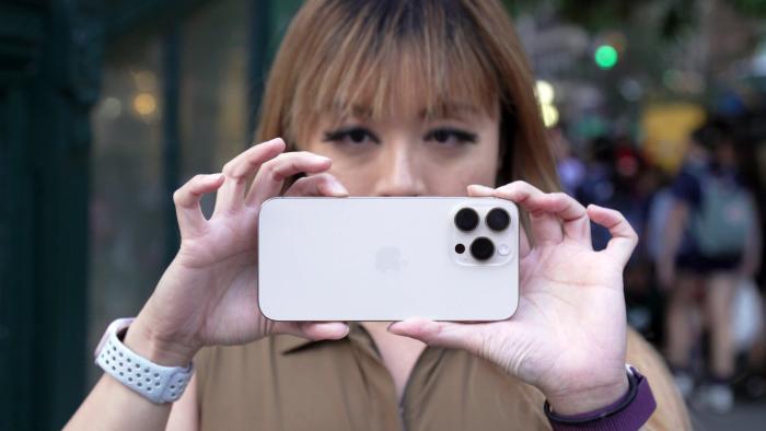 A woman holding the iPhone 16 Pro up in horizontal orientation, with one hand cradling the right side and her other hand positioned with her index finger on the camera control and her thumb at the bottom.